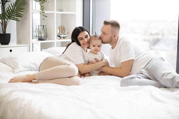 Happy affectionate family lying on king bed and hugging little cutie in white splendid room with city panorama view. Young father kissing sweet daughter in head while mother gently touching. - Foto, imagen