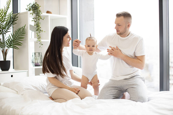 Loving mom and dad holding hands of active girl and helping baby to take first steps on large white bed. Cheerful laughing married couple in casual outfit playing with joyful child. - 写真・画像