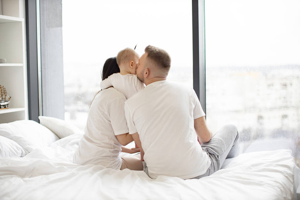 Rear view of loving father kissing his baby daughter while little girl hugging caring mom in morning after waking up. Young married couple with kid resting on white blanket in bedroom of modern flat. - Fotografie, Obrázek