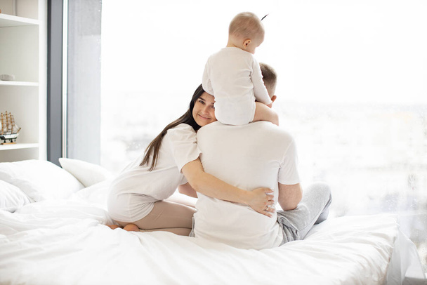 Beautiful brunette wife embracing beloved husband with small kid sitting on dads shoulders and looking out of picture window of airy bedroom. Charming mother showing affection for her family at home. - Фото, изображение