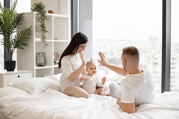 Relaxed young parents giving high five to each other while baby daughter learning new gesture in bedroom interior. Charming mature adults and curious kid making most of leisure time at home. - Zdjęcie, obraz