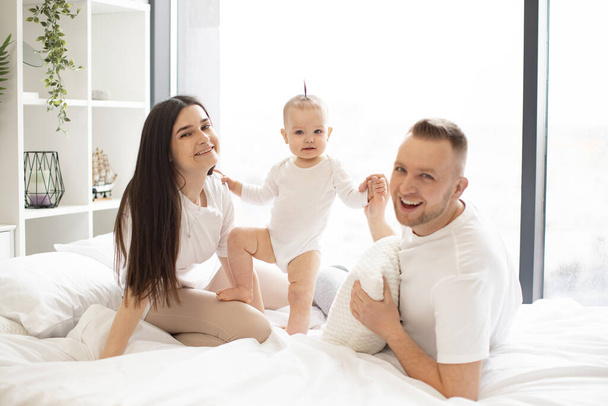 Smiling young father lying on bed while supporting cute baby daughter standing on soft blanket near cheerful mother in casual clothes. Joyful adults and little girl spending family time at home. - Fotografie, Obrázek