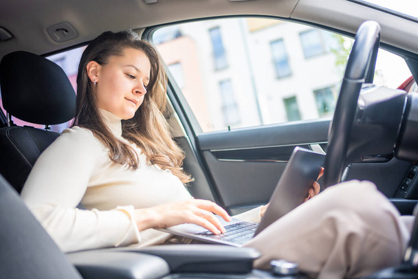 Young businesswoman working on laptop sitting in a car behind the wheel, car work place. High quality photo - 写真・画像