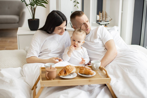 Sweet little baby drinking from sippy cup while joyful brunette woman snuggling with blonde male in cozy bedroom with tray on foreground. Caring family spending quality time together at home. - Fotografie, Obrázek