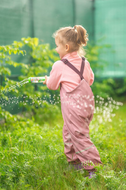 Feliz niña riendo 2-3 años de edad con ropa impermeable tienen una diversión con pistola de agua en el patio trasero del hogar. Foto de alta calidad - Foto, imagen
