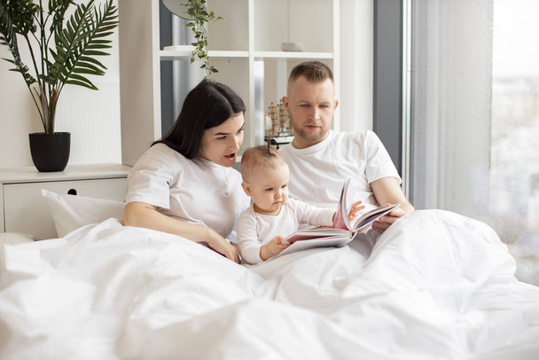 Young married couple in casual outfit sitting in cozy bed with little daughter and reading interactive baby book on Sunday morning. Loving parents making story come alive for curious daughter. - Photo, Image
