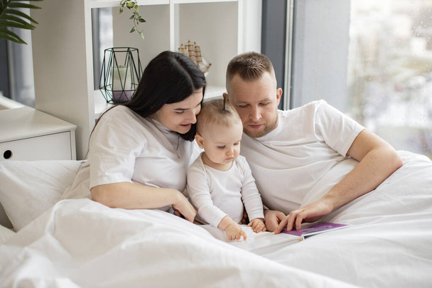 Young married couple in casual outfit sitting in cozy bed with little daughter and reading interactive baby book on Sunday morning. Loving parents making story come alive for curious daughter. - Foto, immagini