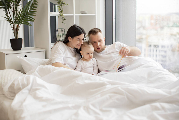 Beautiful woman looking through wordless picture book together with baby girl and attentive male while resting under duvet in bedroom. Mindful parents telling stories to interested kid at weekend. - 写真・画像