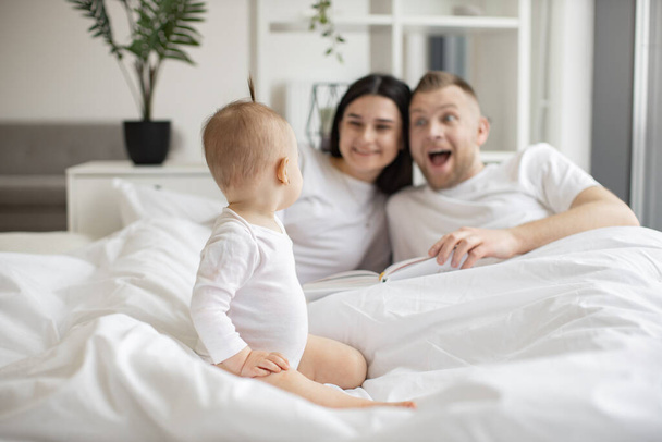 Focus on adorable little child sitting on white soft duvet and watching her parents playing roles of book characters on Sunday morning at home. Concept of family and togetherness. - 写真・画像