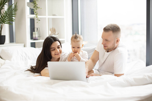 Cheerful married couple in clothes for everyday wear using modern laptop while resting on bed with sweet baby girl during weekend. Young parents surfing internet while little kid eating healthy snack. - Foto, Imagen