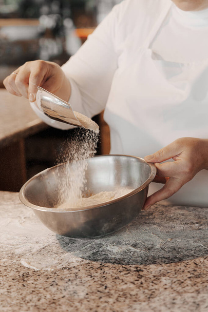 キッチンで生地を準備しながら、金属ボウルにスクープから小麦粉を注ぐ白いエプロンで匿名の女性料理をクロップ - 写真・画像