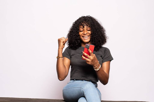 opgewonden mooi jong zwart dame met behulp van haar telefoon vreugde - Foto, afbeelding