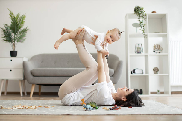 Beautiful brunette woman holding infant girl high in air with arms and legs while lying on carpet in living room. Loving mother letting small daughter fly safely during their funny games. - Fotoğraf, Görsel