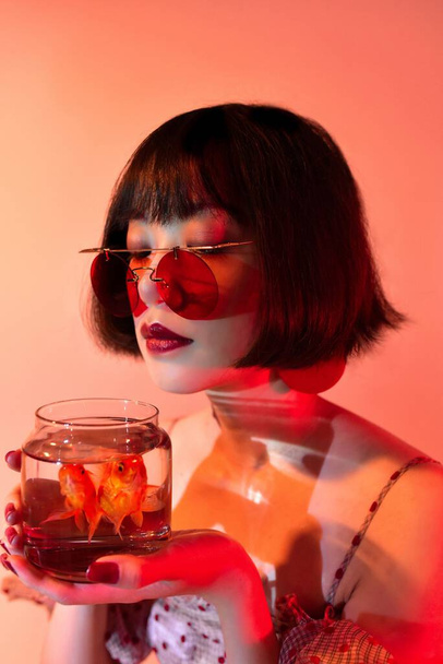 A portrait of a sexy attractive East Asian woman with red sunglasses holding goldfish in a glass and posing - Zdjęcie, obraz