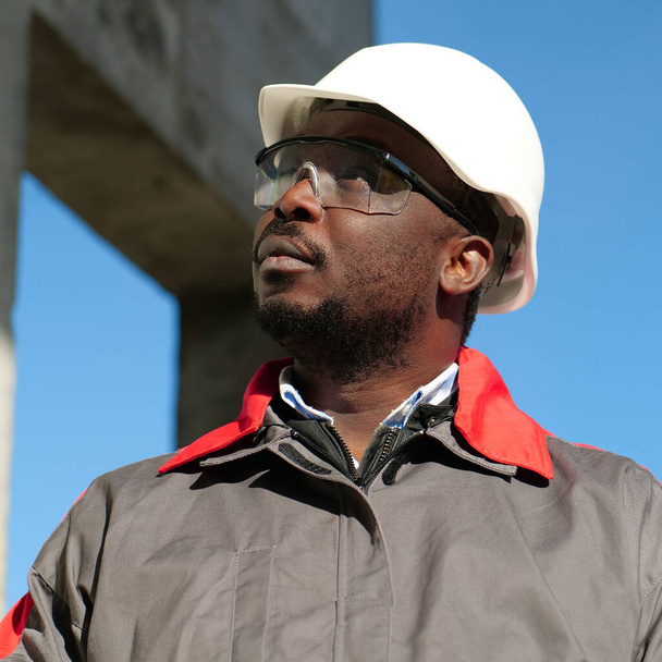 Afrykański amerykański pracownik w twardej czapce i okularach ochronnych stoi na placu budowy i rozgląda się dookoła - Zdjęcie, obraz