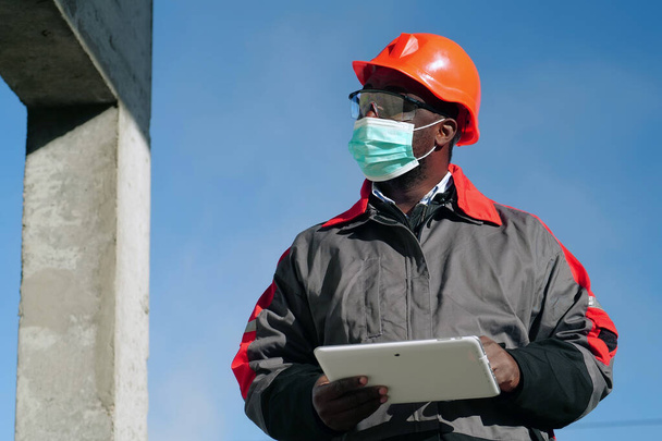 Αφροαμερικανός εργάτης με ιατρική προστατευτική μάσκα στέκεται στο εργοτάξιο με tablet υπολογιστή - Φωτογραφία, εικόνα