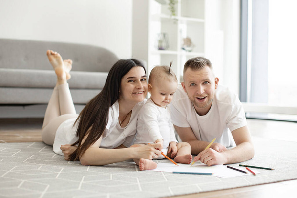 Smiling family of three in casual attire posing with drawing pencils while relaxing on wooden floor in studio apartment. Funny caucasian spouses developing babys imagination using art and colors. - Photo, Image