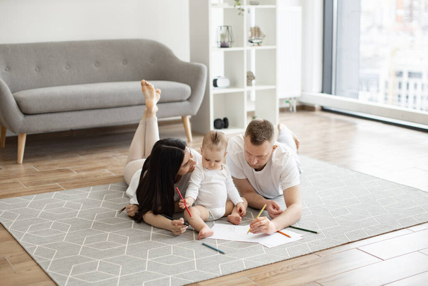 Adorable little baby girl in white cozy bodysuit holding red pencil while sitting on floor with mindful mother and father on both sides. Adult people and kid enjoying first scribbles on paper at home. - Foto, immagini
