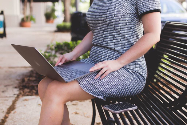 Um close-up de uma fêmea em um vestido casual sentado fora em um banco com seu laptop de joelhos - Foto, Imagem