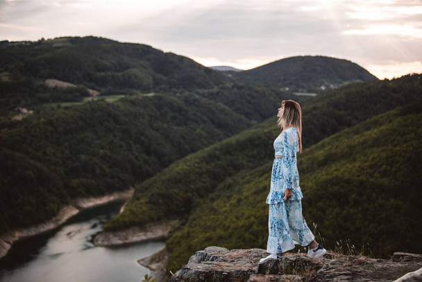 A beautiful Caucasian girl in a dress standing on a rock enjoying the sunset over the river and hills. - 写真・画像