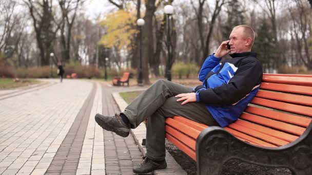 Взрослый мужчина разговаривает по мобильному телефону и улыбается в парке
 - Кадры, видео