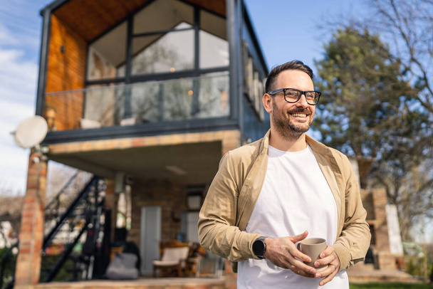A 40-year-old adult man stands confidently in front of his modern home holding a cup of tea or coffee smiling He looks content and happy with his life and proud of his house copy space - Foto, imagen