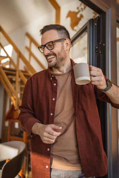 Мужчина, белый 40 лет мужчина носит рубашку стоит на балконе держа чашку чая или кофе улыбаясь счастливым Он наслаждается мирным моментом в его повседневной утренней рутины или на отпуск копировать пространство - Фото, изображение