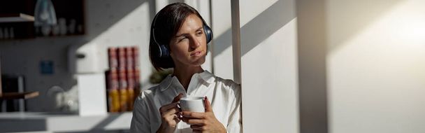European business woman in headphones holding cup of coffee while standing near window at office - Foto, Bild