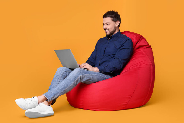 Happy man with laptop sitting in beanbag chair against yellow background - Foto, Imagen