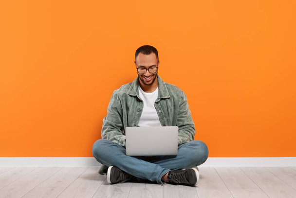 Smiling young man working with laptop near orange wall - Zdjęcie, obraz
