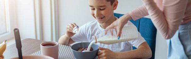 Küçük gülümseyen oğlu, elinde mısır gevreğiyle mutfak masasında oturuyor, annesi süt döküyor. Kahvaltı.. - Fotoğraf, Görsel