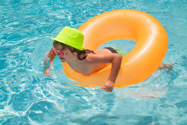 Enfant nage dans la piscine sur anneau gonflable. Nage avec flotteur orange. Jouet aquatique, activité sportive de plein air saine pour les enfants. Mode été enfants en chapeau lunettes de soleil - Photo, image