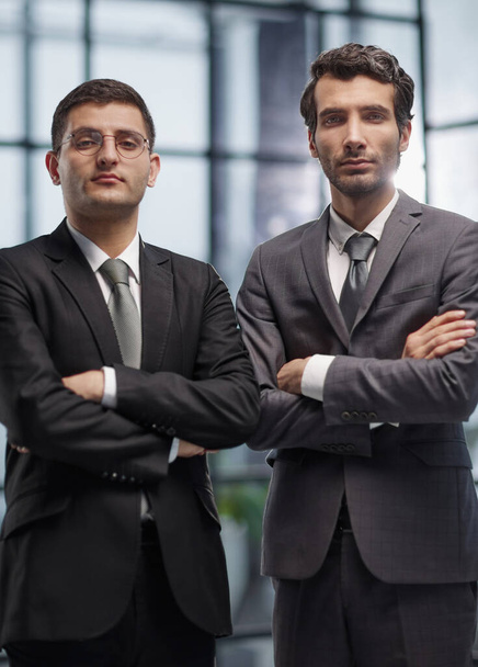 Two serious young businessmen standing with arms crossed in office - Foto, afbeelding