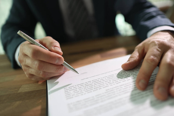 Businessman puts personal signature on contract paper sheet sitting at wooden table. Man holds ball-pen in hand ready to sign document in office - Photo, Image