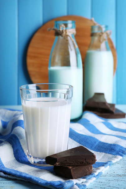Glass and bottles of milk with chocolate chunks on stripped napkin and color wooden planks background - Фото, изображение