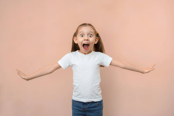 Shocked emotional caucasian girl 7 years old in a white t-shirt on a pink background. - Foto, Imagen