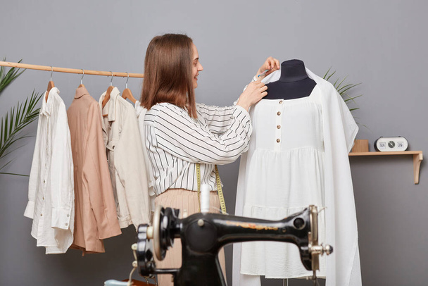 Side view of joyful professional woman dressmaker putting pin in fabric hanging on mannequin while working in modern workshop, creating new collection, sewing dress. - Foto, Bild
