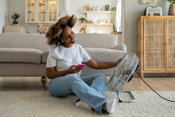 Happy relaxed young African American woman holding smartphone sitting in front of electric fan at home, smiling black girl feeling good while relaxing near ventilator during extreme heat - Foto, Bild