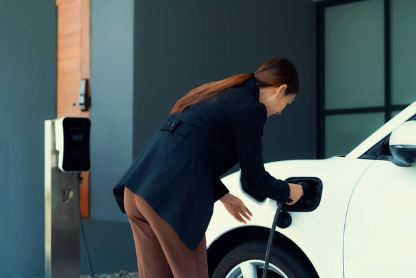 Progressive woman install cable plug to her electric car with home charging station. Concept of the use of electric vehicles in a progressive lifestyle contributes to clean environment. - Photo, Image