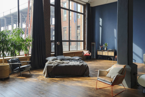 dark interior of a modern stylish huge open-plan loft-style studio apartment with columns and high ceilings. dark blue primed walls are decorated with wood. sunlight enters through huge windows. - Foto, Bild