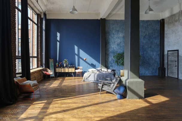dark interior of a modern stylish huge open-plan loft-style studio apartment with columns and high ceilings. dark blue primed walls are decorated with wood. sunlight enters through huge windows. - Фото, изображение