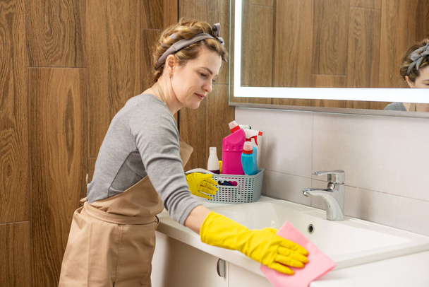 Weekend homework. Young pretty woman cleans in cozy living room at home. A beautiful woman in home clothes and rubber gloves in a bright room - 写真・画像