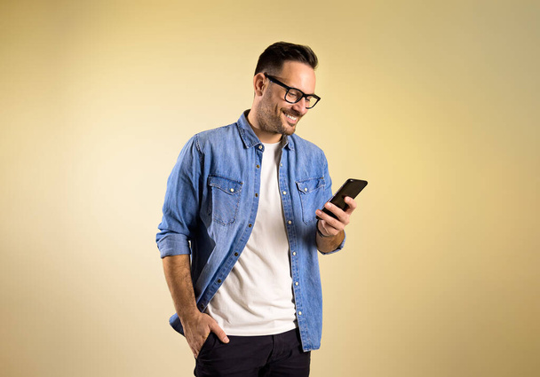 Charming male entrepreneur with hand in pocket smiling and messaging over mobile phone. Happy young man wearing denim shirt and eyeglasses standing isolated on yellow background - Photo, Image