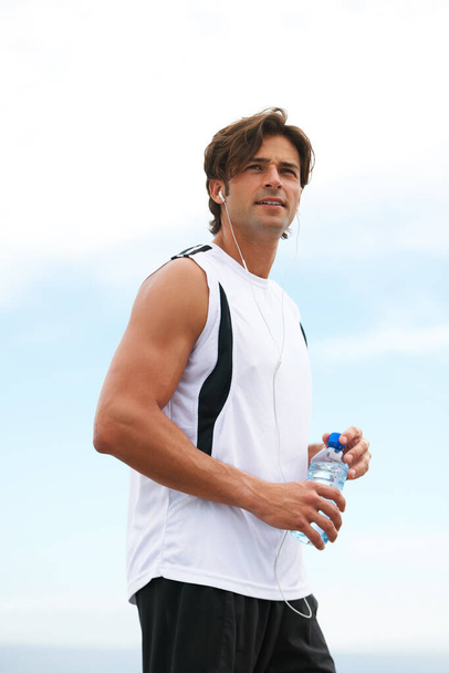 Él tiene una fuerte motivación para la aptitud física. Hombre joven guapo sosteniendo una botella de agua, escuchando música y mirando hacia otro lado después de su carrera matutina - Foto, imagen