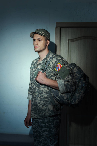 Soldado americano disgustado en camuflaje de pie en el pasillo en casa por la noche  - Foto, imagen