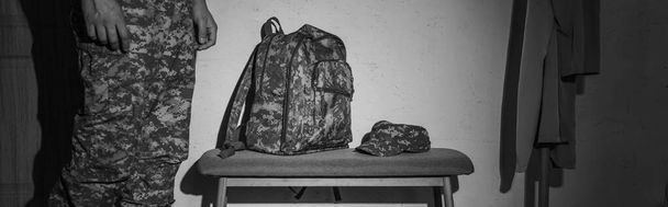 Foto en blanco y negro del soldado en uniforme de pie cerca de la mochila y el coche en el pasillo en casa, pancarta  - Foto, imagen