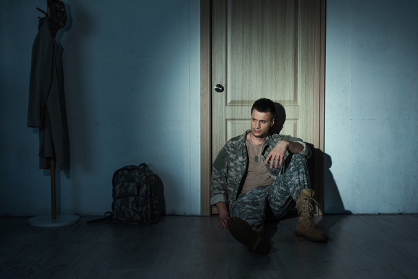 Разочарованный военный ветеран в форме сидит на полу рядом с дверью в коридоре ночью  - Фото, изображение