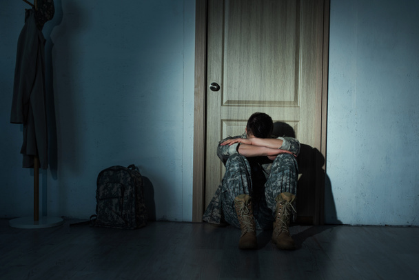 Одинокий солдат в форме сидит ночью возле рюкзака и двери дома  - Фото, изображение