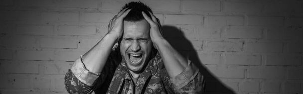 Black and white photo of stressed military veteran screaming while suffering from post traumatic stress disorder at home, banner  - Photo, Image