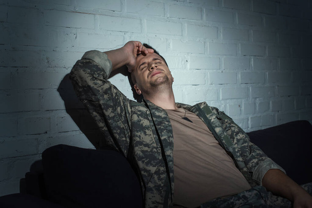 Militärangehöriger mit psychischer Störung berührt Kopf, während er nachts auf Couch zu Hause sitzt  - Foto, Bild